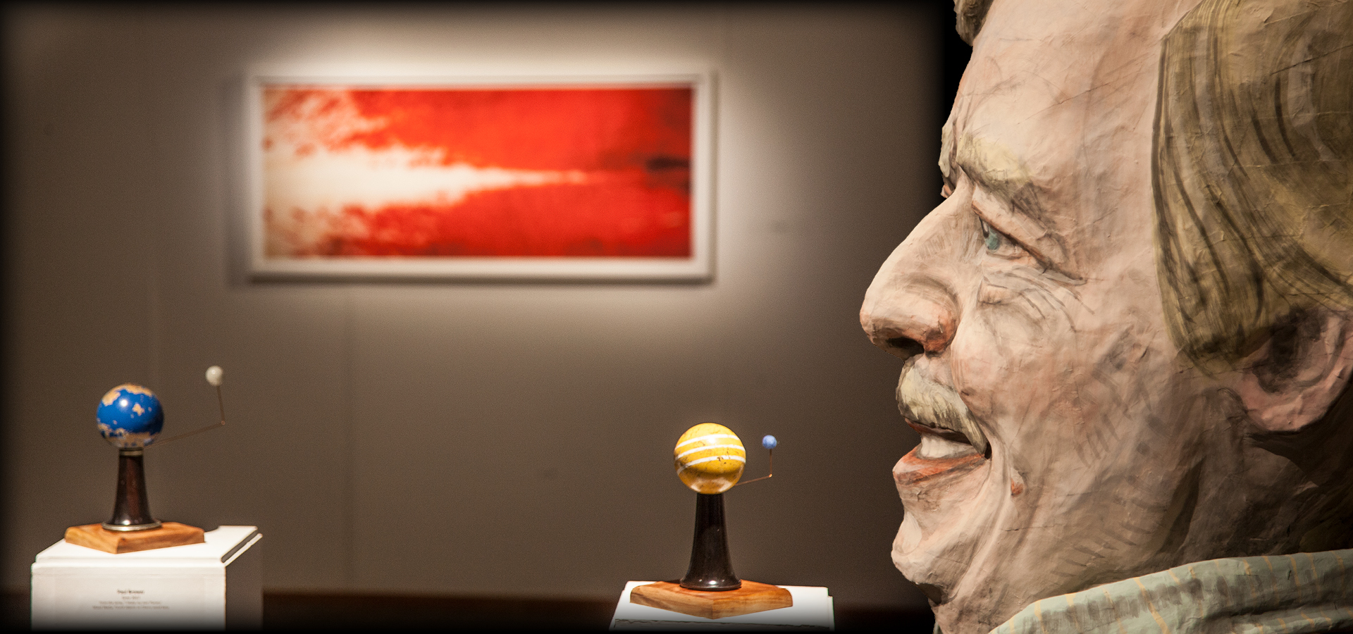 A larger-than-life sculpture of a head in front and to the side of an abstract, blurred painting hung on a gallery wall. In front of the painting two small sculptures stand on pedestals, acting as diagrams of moons orbiting around planets 