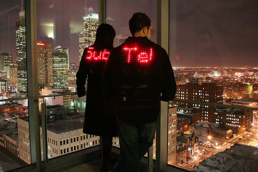 Two people stand at the corner window of a room, overlooking a night city skyline. Both people are wearing dark jackets with words spelled out in L.E.D. lights on the back: one says "Sub" and the other says "Tel".