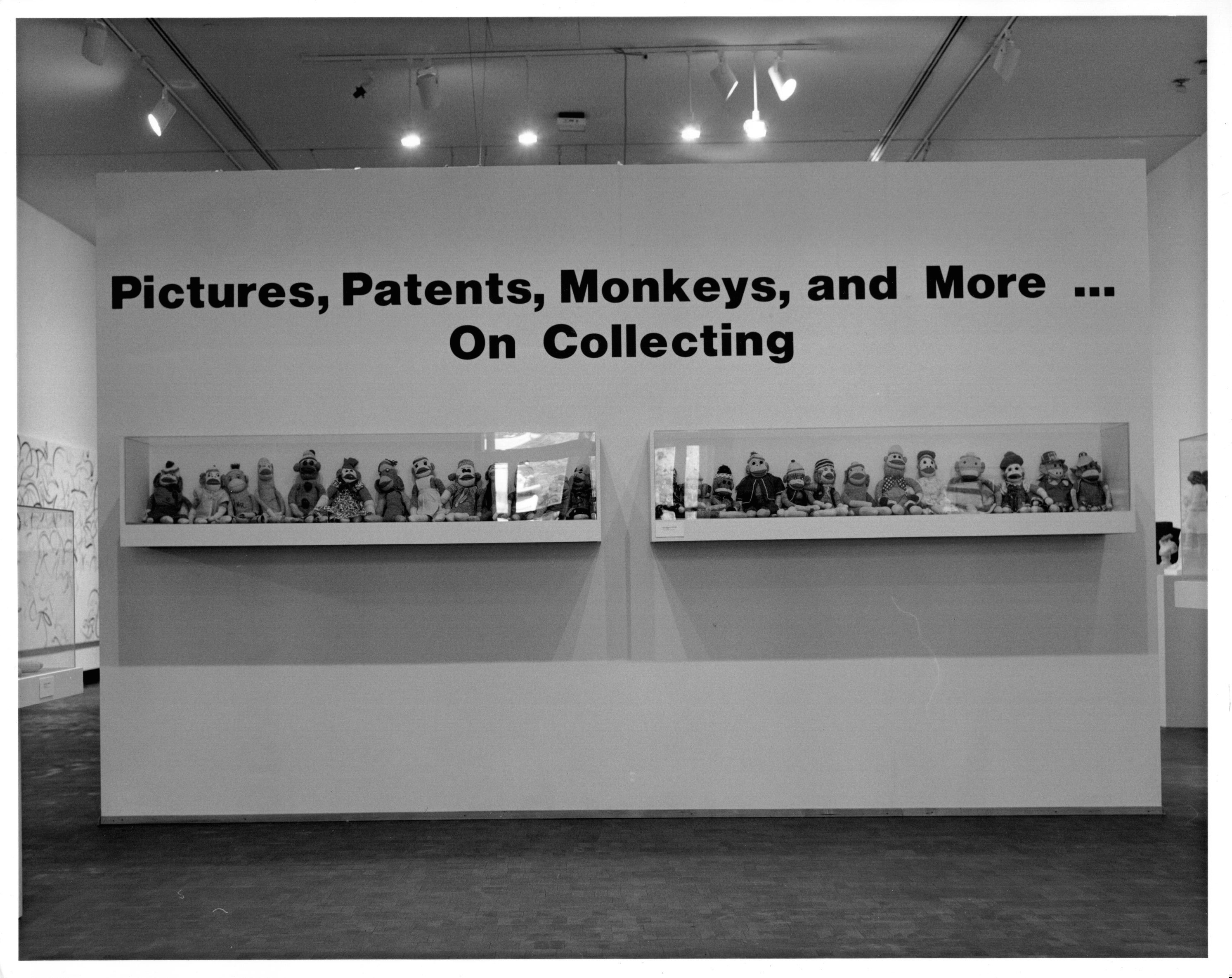 A photograph of a temporary wall that fills up the entire frame. The light work from behind this wall can be seen at the top of the frame. At the top of this wall it reads in large bold black letters &quot;Pictures, Patents, Monkeys, and More ... On Collecting