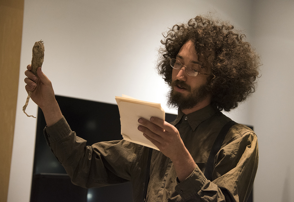Robert Yerachmiel Snyderman, sporting a curly afro, glasses, beard and button-up shirt, reads from a handful of notepaper while holding up what appears to be a wrapped bundle of sage