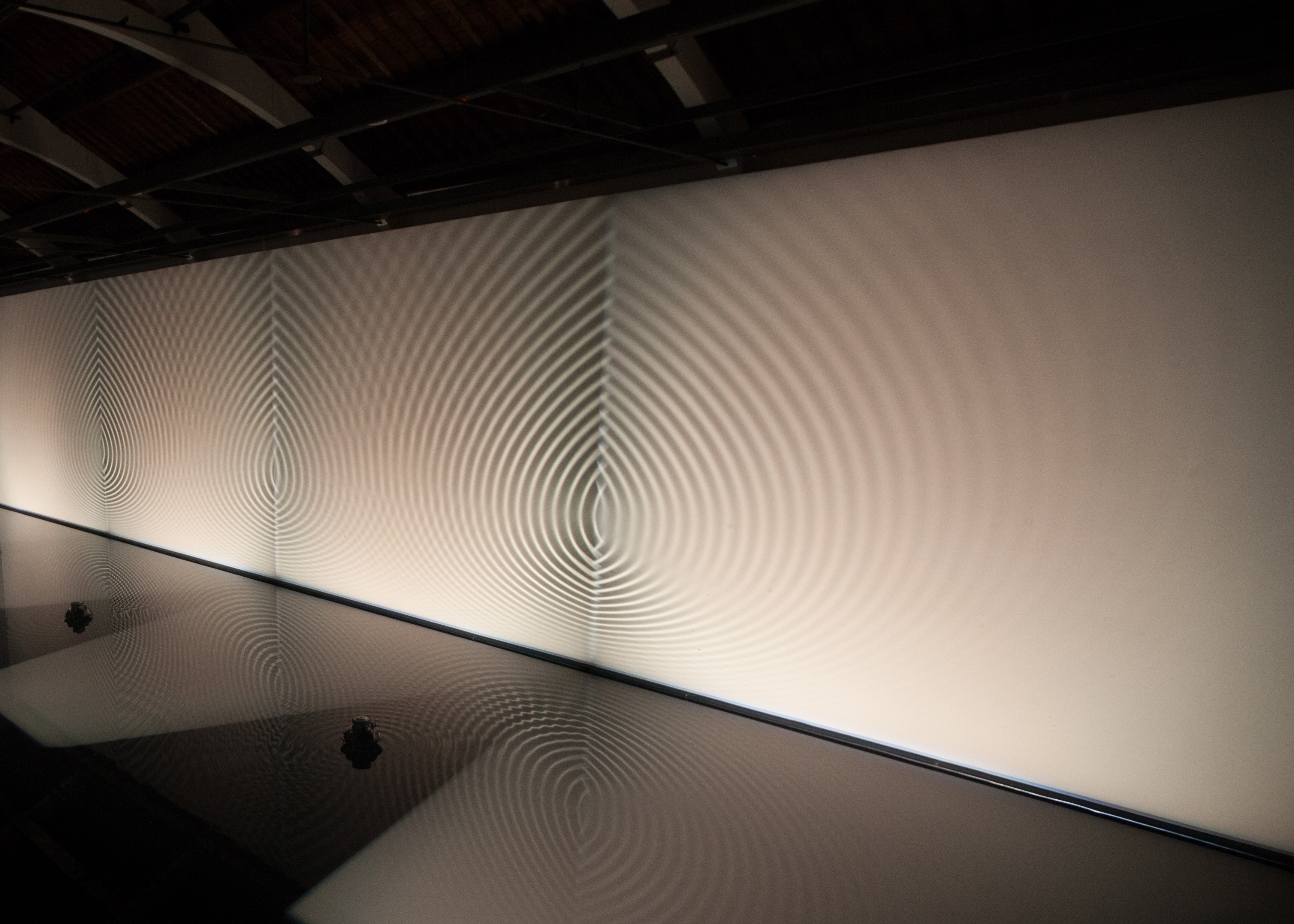 a shallow pool rippled by sound waves bounces reflected light onto a long blank wall