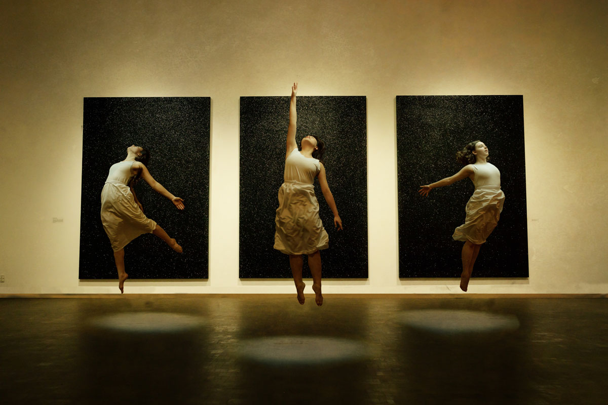 three dancers leap upward, backs arched. Center dancer reaching. Black paintings behind frame each.