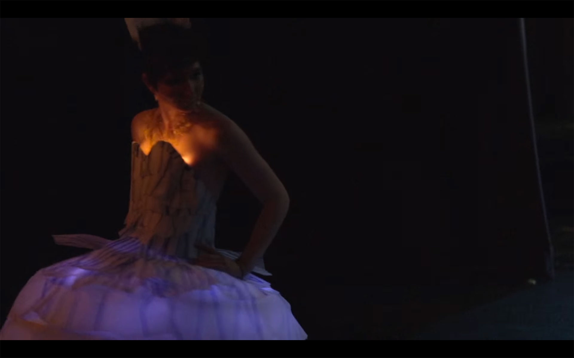 very dark scene. a figure in a poofy white dress is illuminated dimly by lights within the dress.