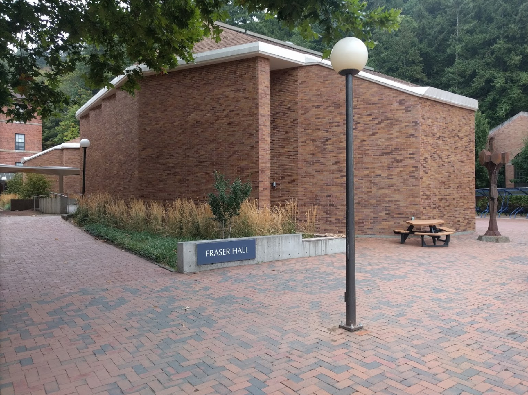 A brick plaza surrounds a blocky brick building with angled corners. A sign reads Fraser Hall. In the background is a fir forest, more brick buildings, a bike rack and an outdoor sculpture.