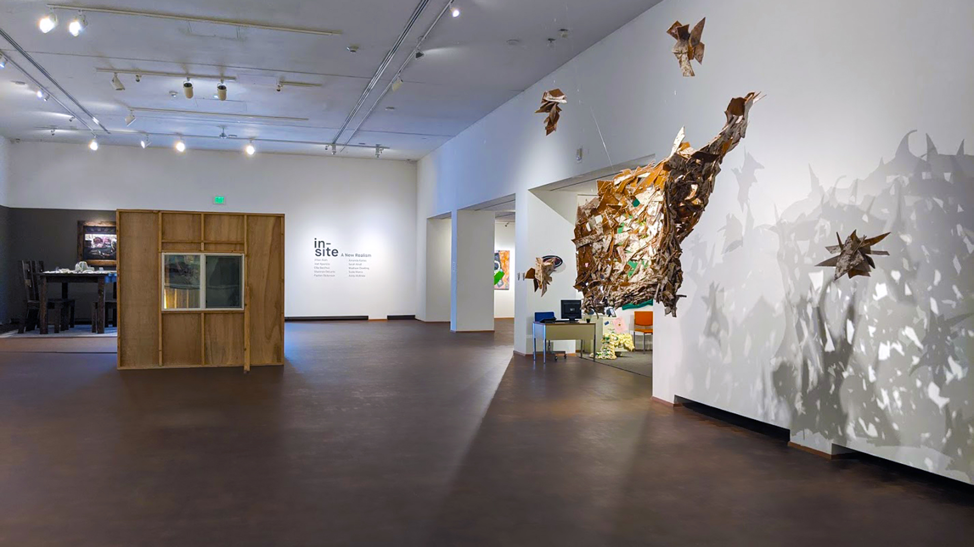 An exhibition inside the Western Gallery: To the right, an abstract 3D artwork hangs from the gallery ceiling, casting interlaced patterned shadows on the wall. To the left, an oversized dining table sits within temporary walls built to look like a room within the gallery.