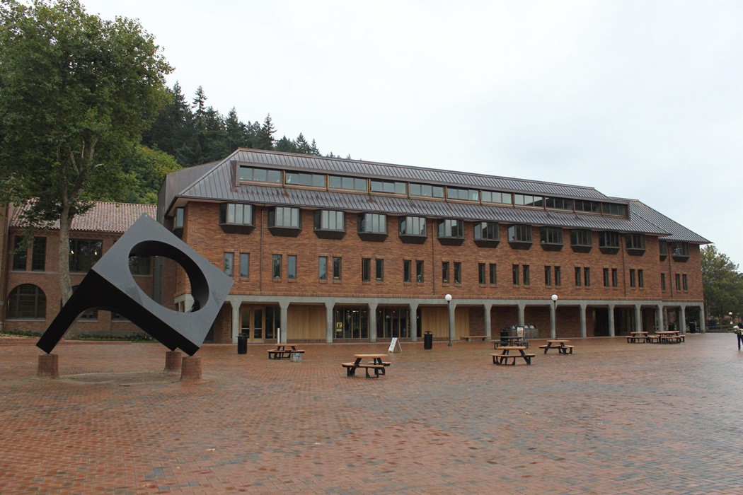 A brick plaza spans before a long, 4 story brick building with metal roof, lots of windows, and covered walkway on the exterior ground floor. To the left stands a modern metal outdoor sculpture.