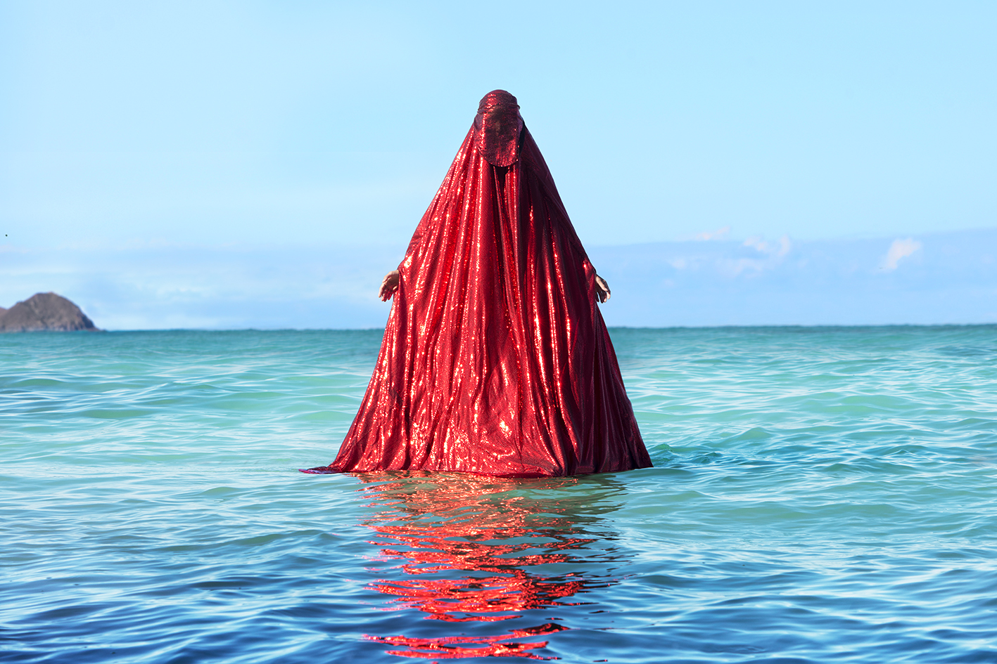 a figure fully covered in a bright, sparkling red chador emerges from blue ocean water. The red chador reflects upon the ripples of the water.