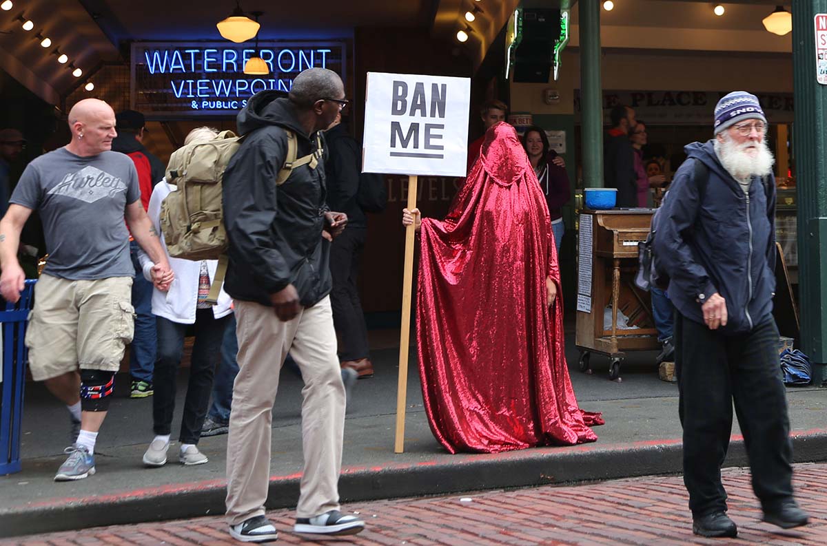 a figure in a red chador stands in a Seattle market holding a sign reading "ban me" while bemused onlookers pass by