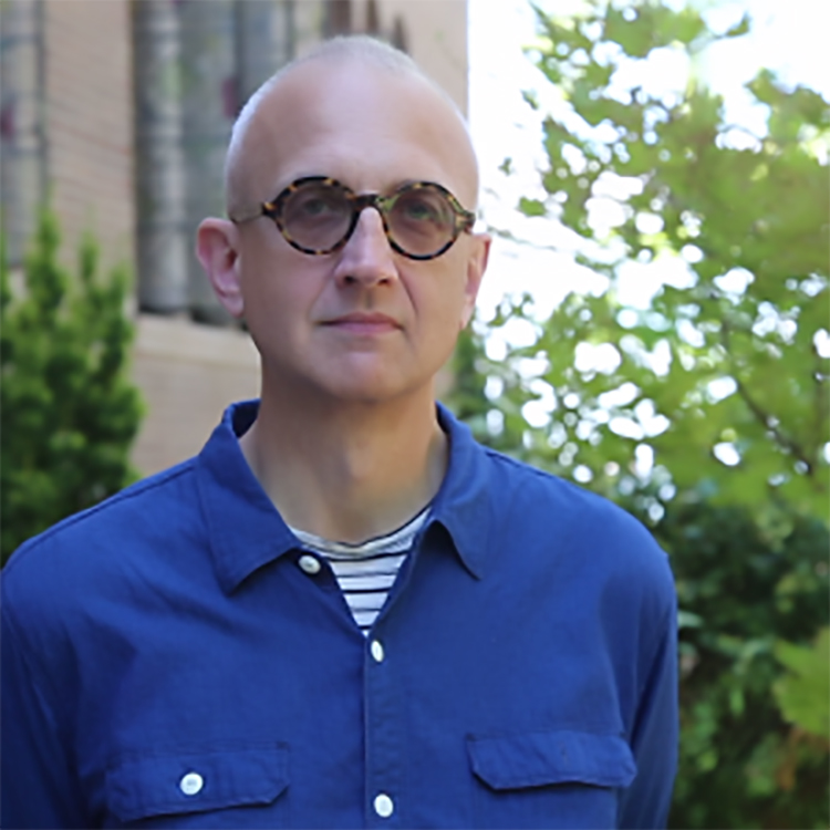 a person with buzzed haircut, blue shirt and slightly tinted glasses standing outdoors