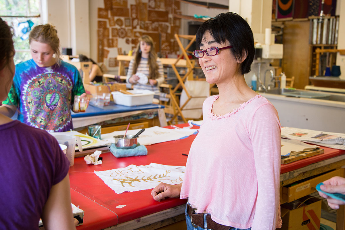a woman in a pink blouse smiles at a person off camera. The woman is standing in a fiber arts studio.
