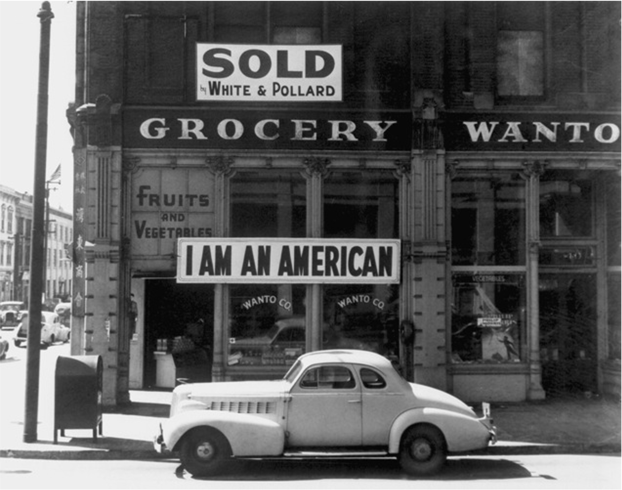 a 1930s coupe automobile sits in front of a grocer with a sign above that says "I am an American" and another that says "sold"