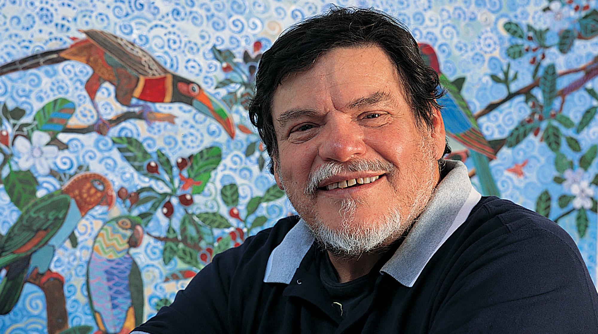 A friendly looking person with a gray beard smiles in front of art depicting parrots