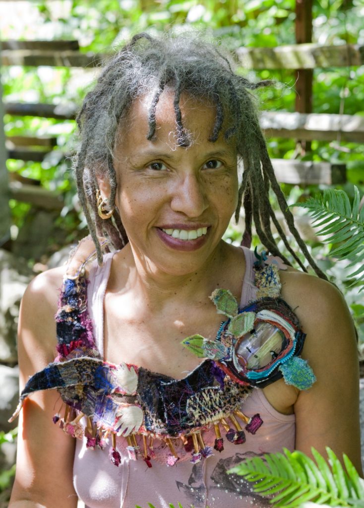 A woman with an eclectic necklace sits among ferns  with a bright smile