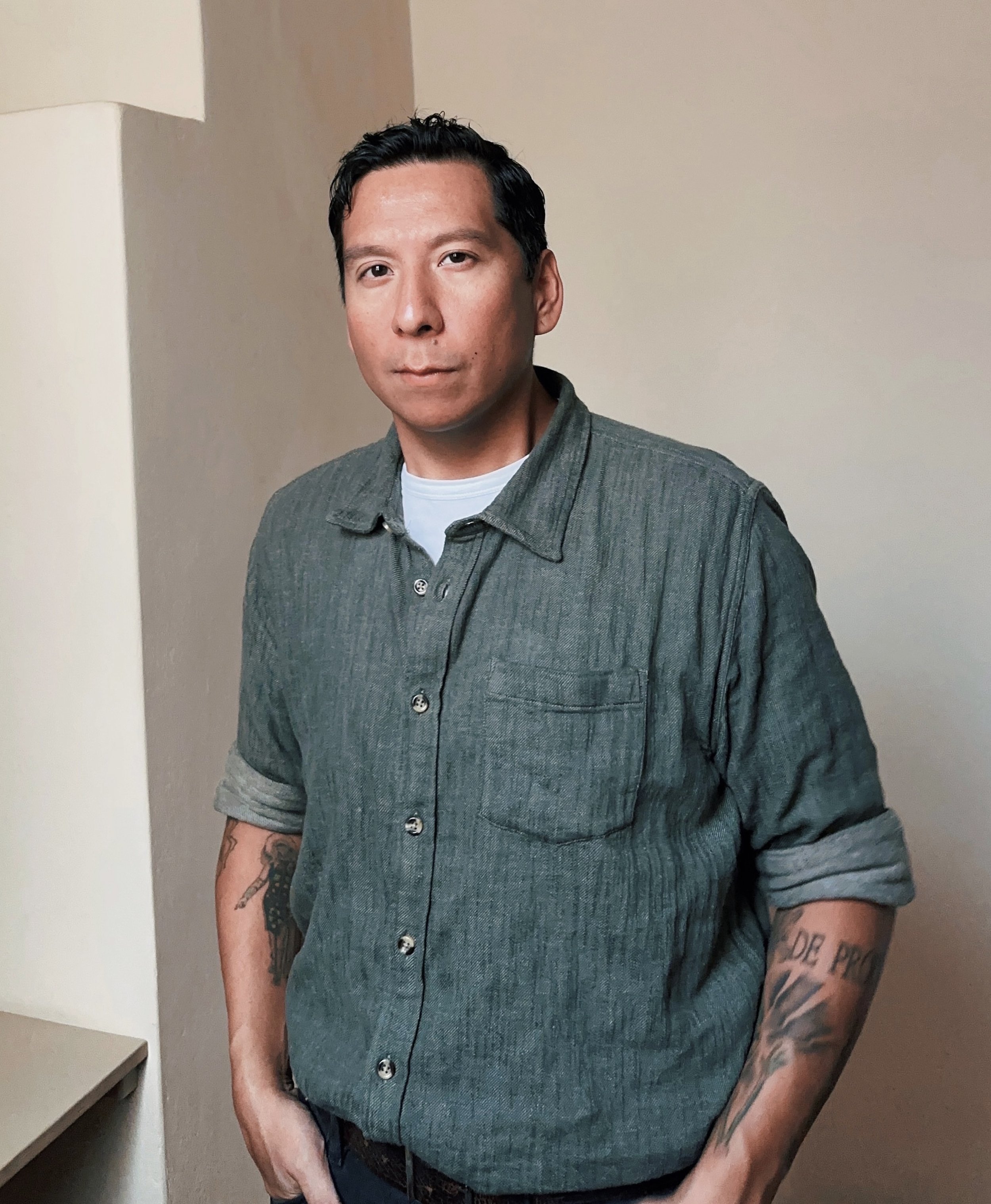 Sky Hopinka standing relaxed in a corner with hands in pockets. His sleeves are rolled up, showing indigenous-themed tattoos on his forearms.