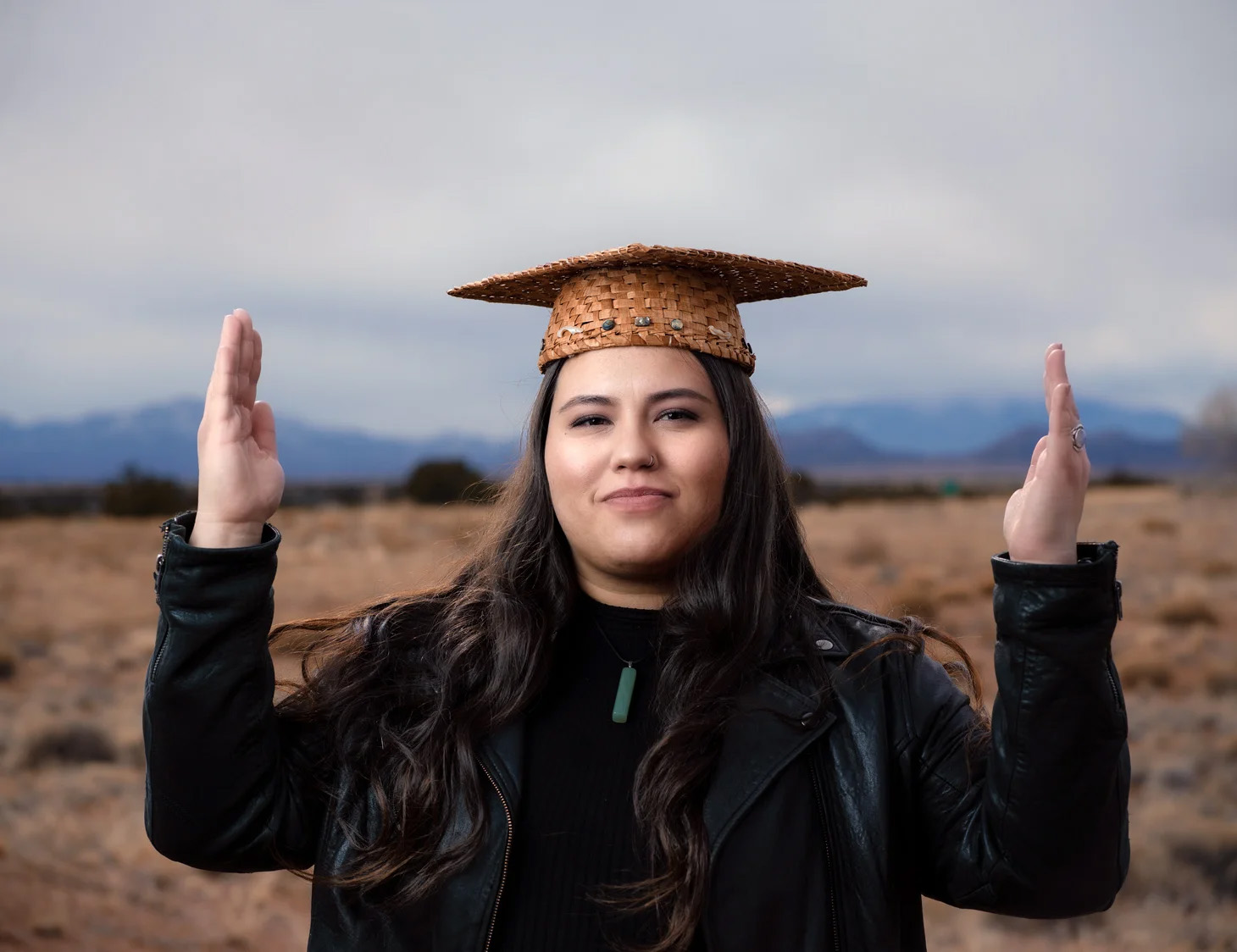 a young person with straight black hair wears a basket-weave mortarboard hat and holds their arms out with hands up and elbows at right angles. Their hands face inwards toward their head.