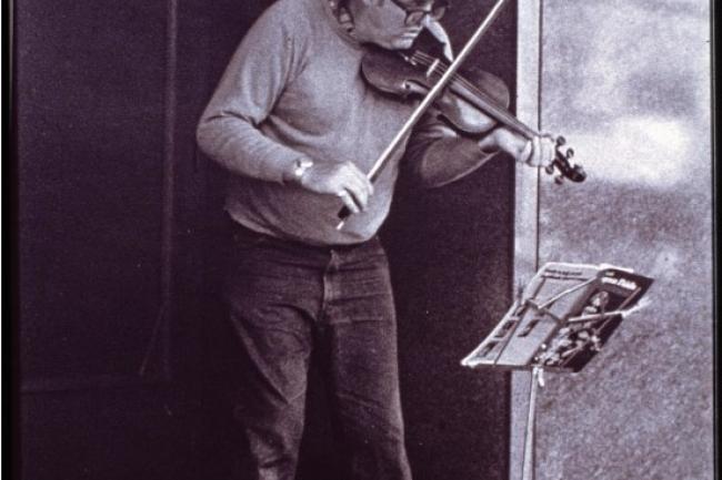 A violinist playing against a building corner, focused on a music stand in front of them, an open violin case on the ground below.