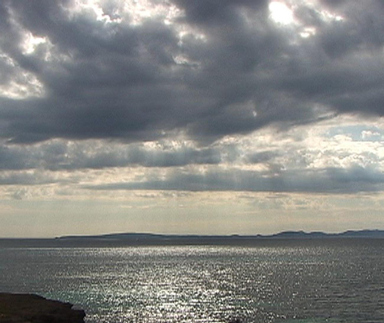 sunrays shine down from gray clouds onto a bumpy ocean. Silhouetted islands appear on the horizon line.