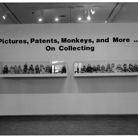 A photograph of a temporary wall that fills up the entire frame. The light work from behind this wall can be seen at the top of the frame. At the top of this wall it reads in large bold black letters &quot;Pictures, Patents, Monkeys, and More ... On Collecting