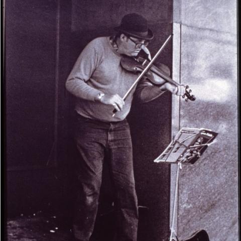 A violinist playing against a building corner, focused on a music stand in front of them, an open violin case on the ground below.