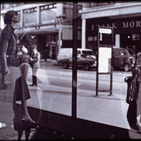 A model or manequin faces the street from inside a store window. A saxophonist leans back and plays facing the shop window.