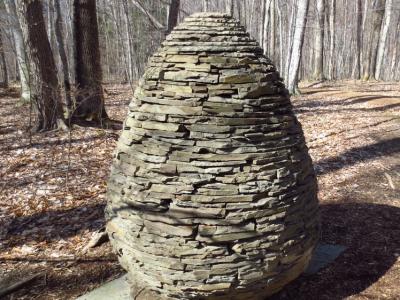 a person-sized object that looks like a beehive or an egg made of piled flat stones sits in a forest of bare-limbed deciduous trees.