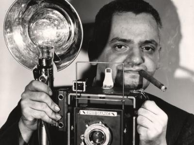 person with a lit cigar in their mouth holding a large older medium format camera with a reflector and flashbulb 