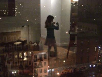 A person in their living room reflected on a window which looks out over a city at night