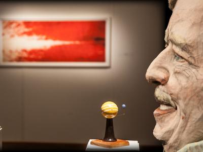 A larger-than-life sculpture of a head in front and to the side of an abstract, blurred painting hung on a gallery wall. In front of the painting two small sculptures stand on pedestals, acting as diagrams of moons orbiting around planets 