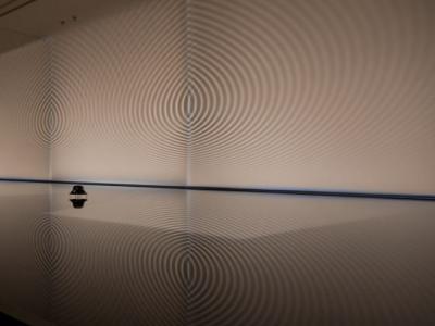 ripples of light and shadow radiate outward from two points on a wall, spanning the entire wall and intersecting between the two points. This also reflects onto the clean floor in front of the wall.