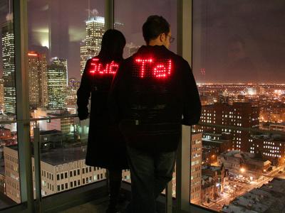 Two people stand at the corner window of a room, overlooking a night city skyline. Both people are wearing dark jackets with words spelled out in L.E.D. lights on the back: one says "Sub" and the other says "Tel".
