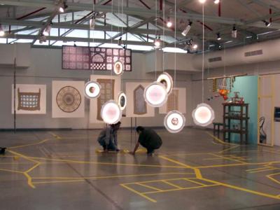 artists installing work in a gallery. glowing discs hang from the beams of the ceiling.