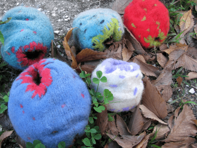 five balls of colorful felted yarn lay in a patch of gravel, leaves, and grass