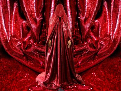 a figure fully covered in a bright, sparkling red chador stands in the middle of the image. The backdrop of the image, and ground under the figure is the same red chador viewed close enough to see the individual sequins it is made of.