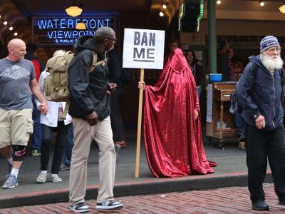 a figure in a red chador stands in a Seattle market holding a sign reading "ban me" while bemused onlookers pass by