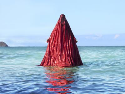 a figure fully covered in a bright, sparkling red chador emerges from blue ocean water. The red chador reflects upon the ripples of the water.