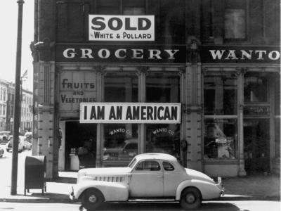 a 1930s coupe automobile sits in front of a grocer with a sign above that says "I am an American" and another that says "sold"