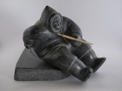 carved stone sculpture of an indigenous hunter sitting on a platform, holding a long wood tool tied to a rope