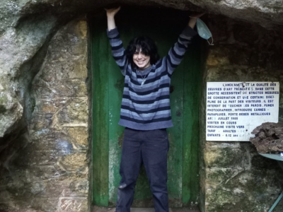 a person in a striped shirt poses with arms up inside the opening to a cave or mine with a green door.