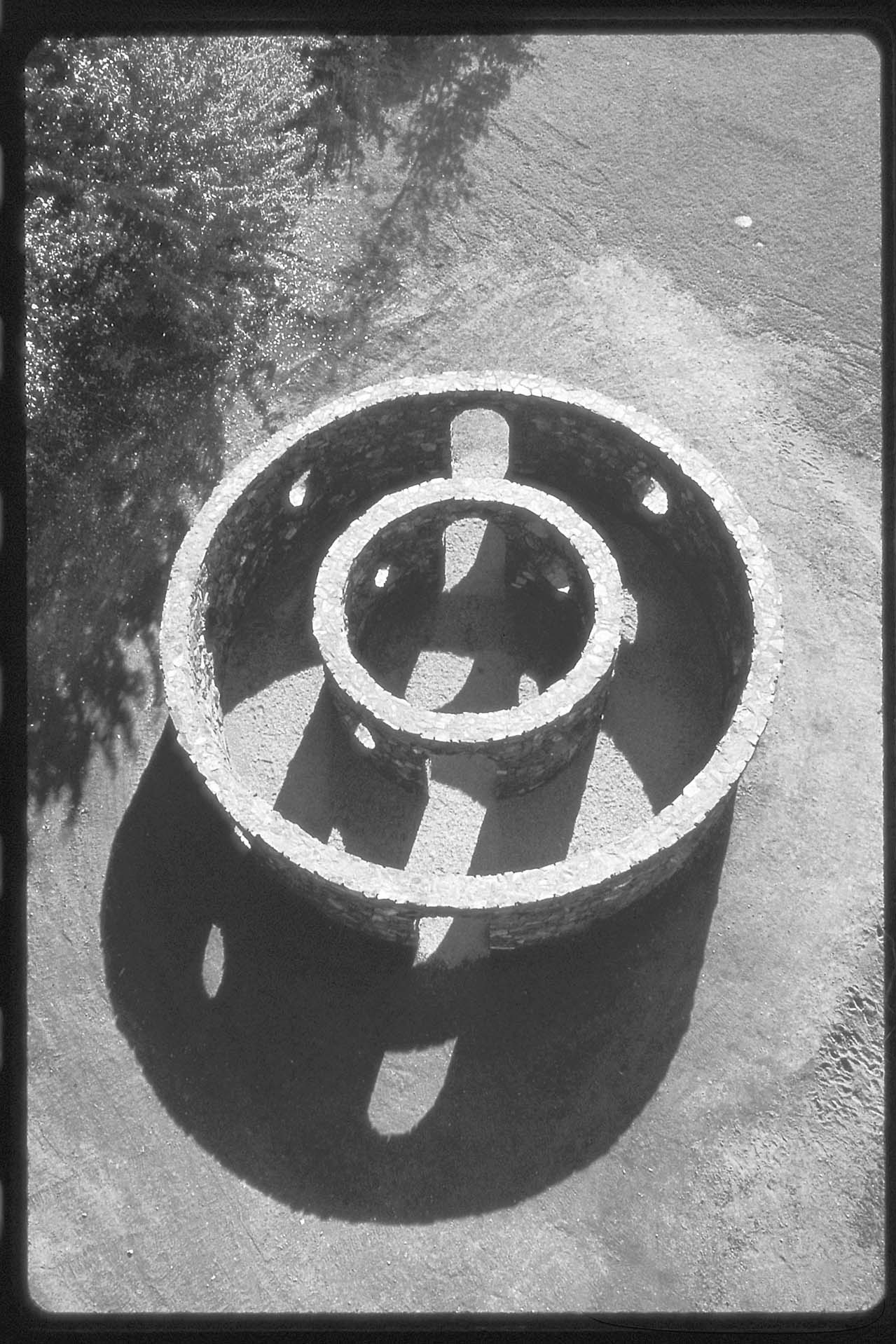 aerial view of two concentric rings of masonry. angled light reveals shadows of circular openings on the ground adjacent. Black and white photo.