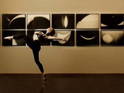 a dancer balances on one foot - perhaps in the midst of a giant step or leap. On the wall behind is a series of monochromatic images of curvilinear light and dark forms.